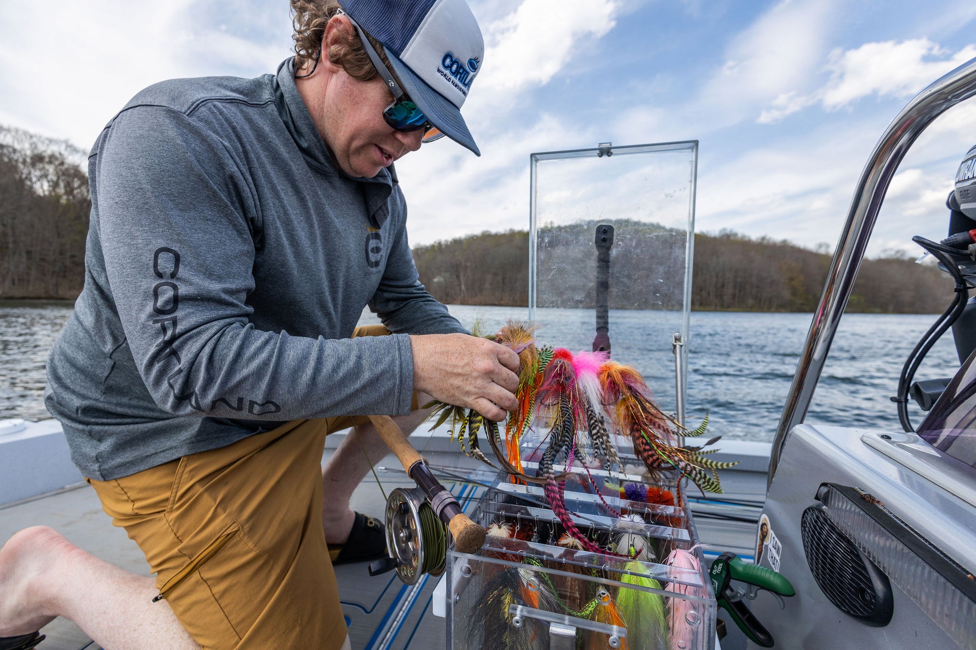 A man is kneeling down on his bloat and going through various fly fishing lures