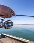 A hand is holding onto a fishing pole that is lined with Cortland Master Braid Bronze. There is a lure at the end of the line and water in the background.