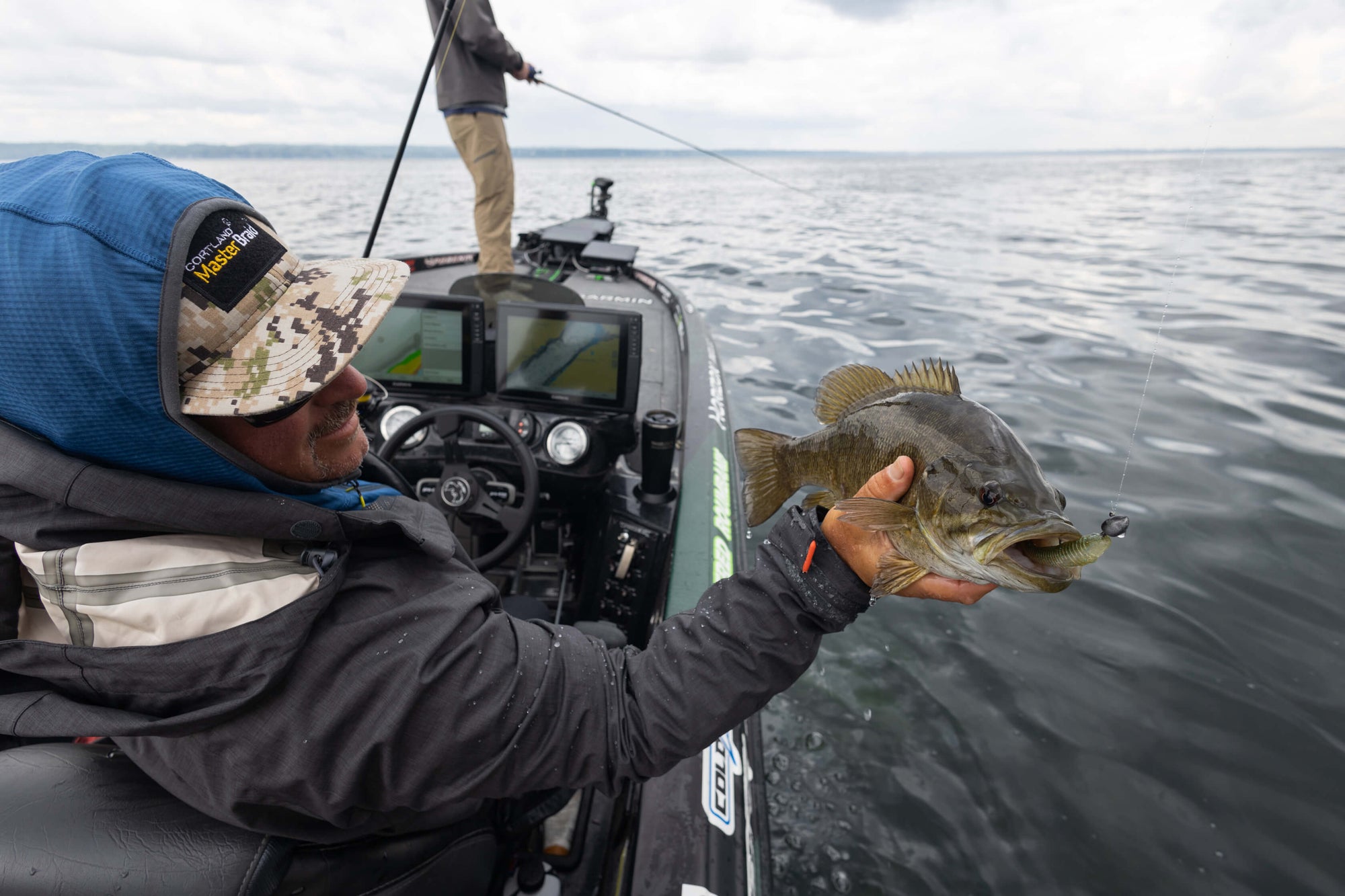 There is an angler sitting on a boat and holding up a fish. And another individual fishing at the bow of the boat. 