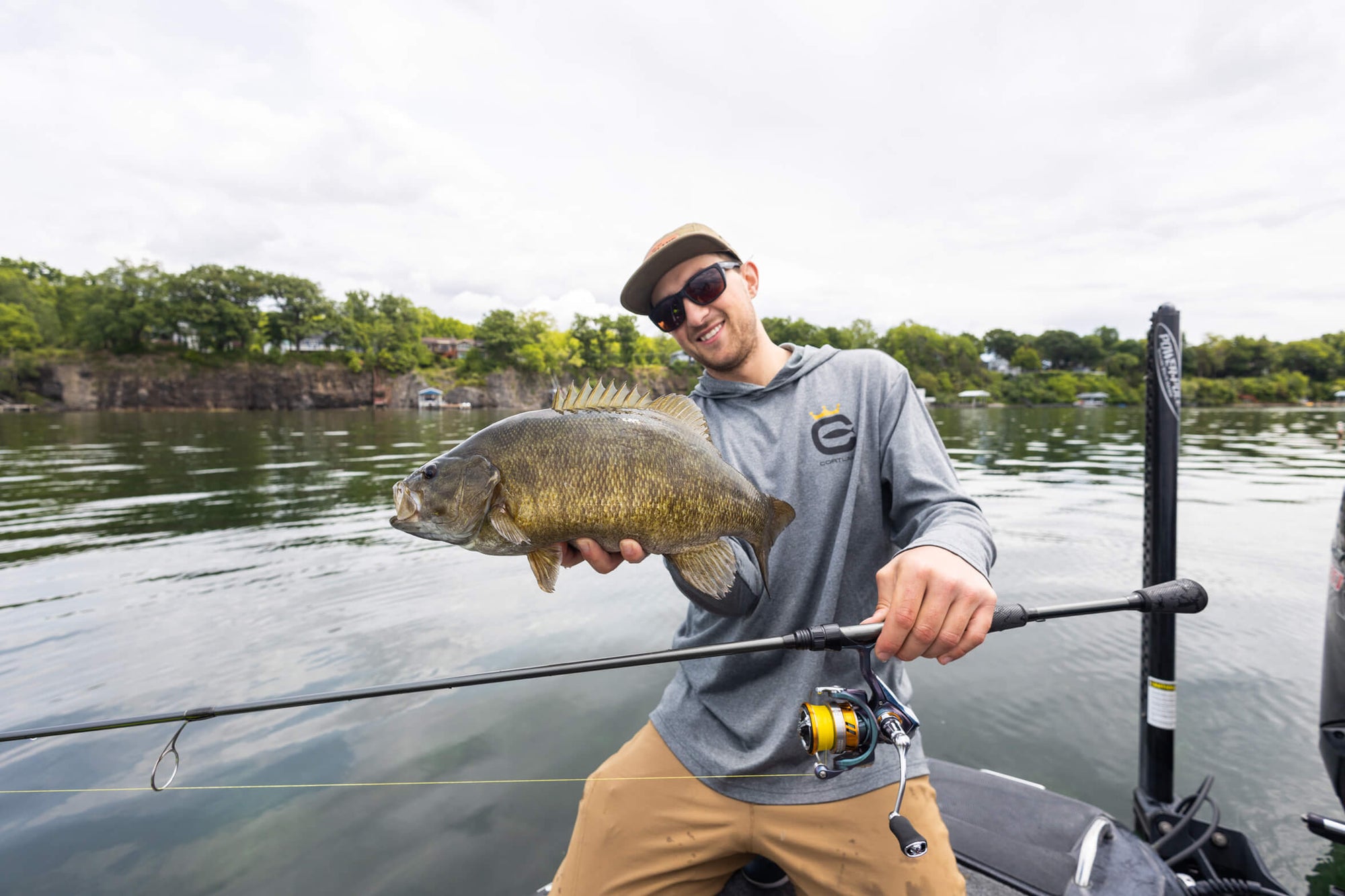 A man is smiling while holding up a fish and his fishing rod. 