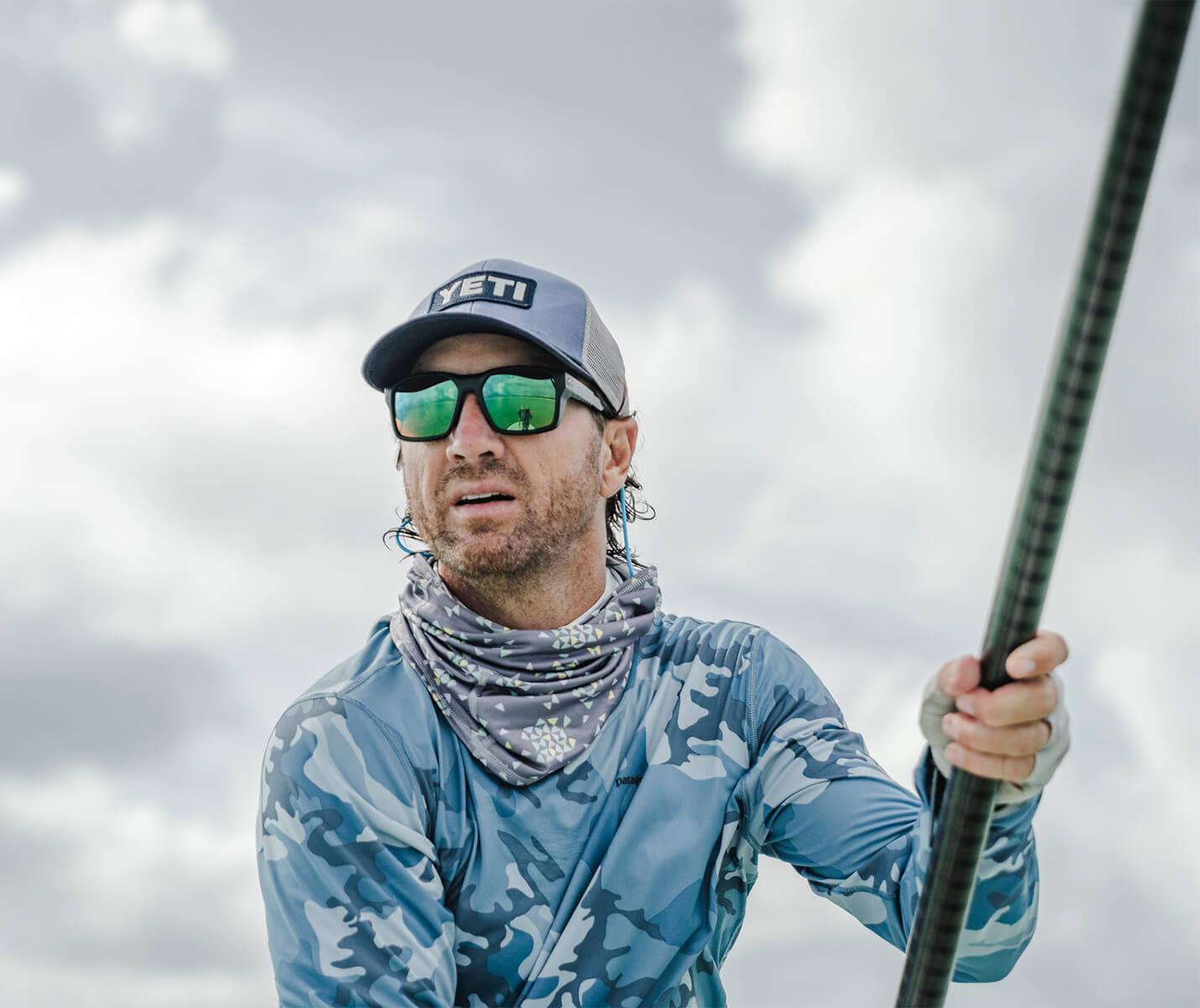Keith is wearing sunglasses, a hat, and a blue cameo shirt. He is looking out in front of him, while holding up a pole