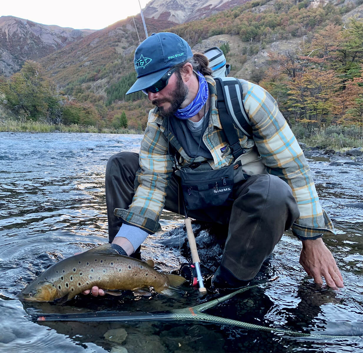 Lance Wilt kneeing down with a fish in his hand. There is a fly rod in his lap and a fishing net in the river.