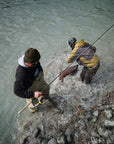 Two individuals are standing in a river. One is watching while the other is pulling in their fish.