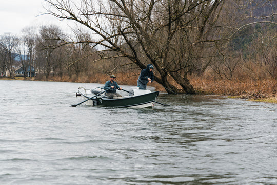 Two anglers are fishing on a boat. One is reeling in his fly line, while the other is paddling 