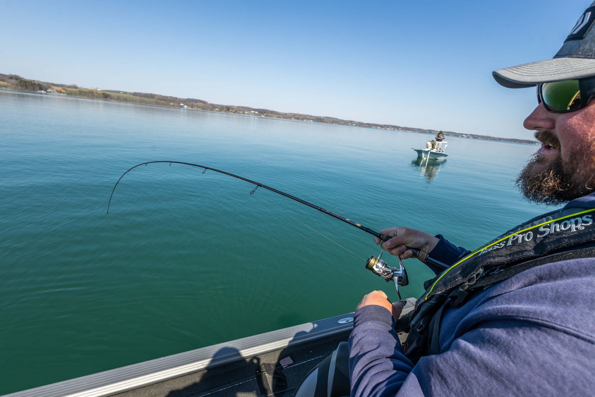 A man is overlooking the water, as he is reeling in his pole. There is another person standing on a boat in the background. 