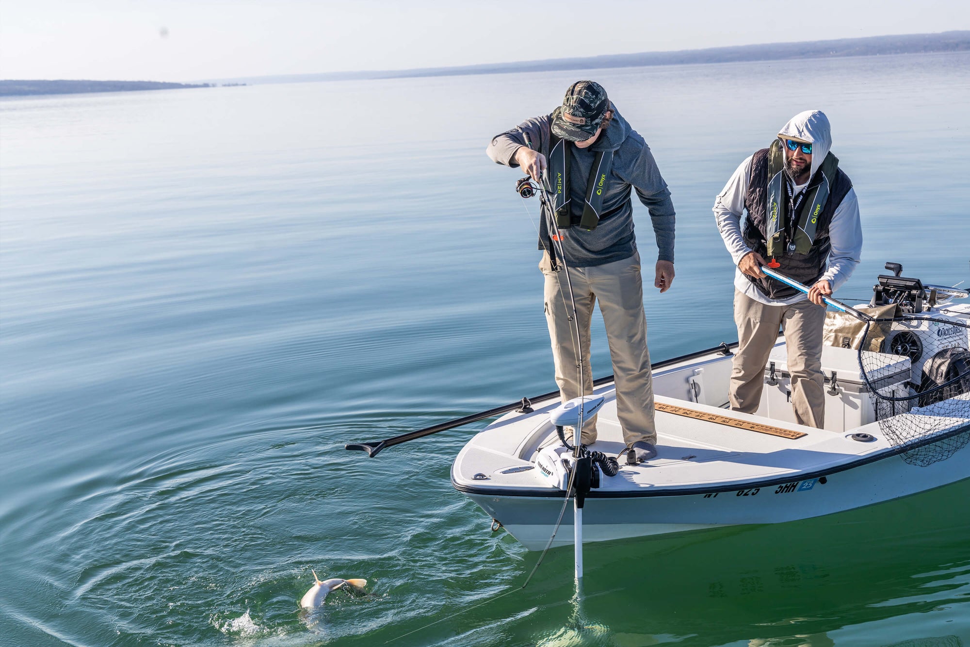 Two anglers are standing on a boat. One has prepared the net, while the other is reeling in the fish he caught. 