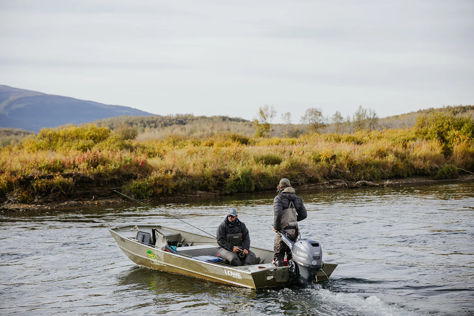 Two people fishing from a boat on a river with mountains in the background