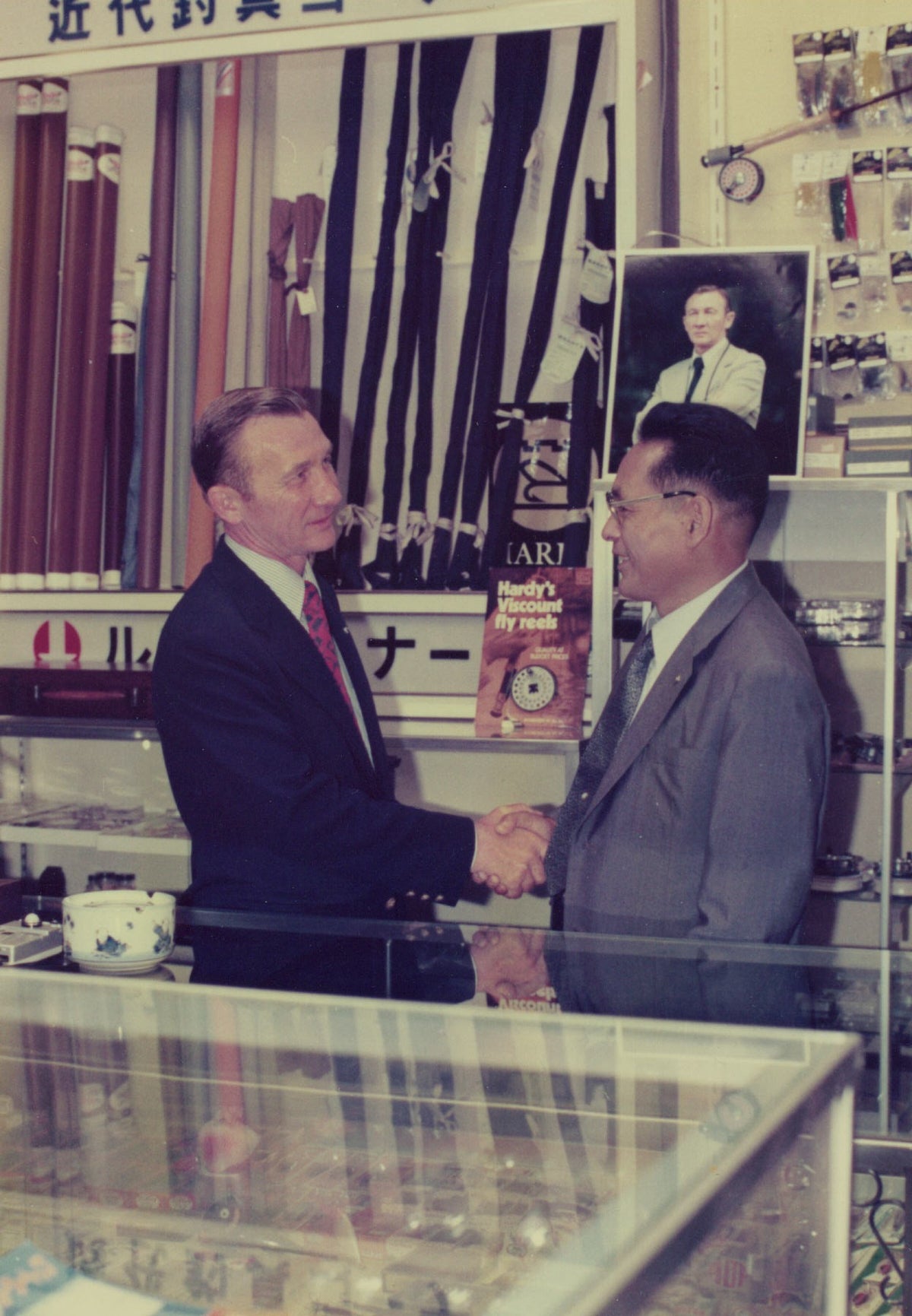 Two men shaking hands behind a counter in a store with various items on shelves.