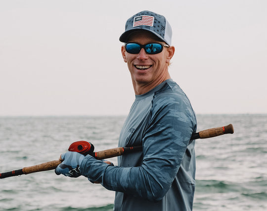 Spencer holding up a fly fishing rod and smiling at the camera. There is a body of water in the background