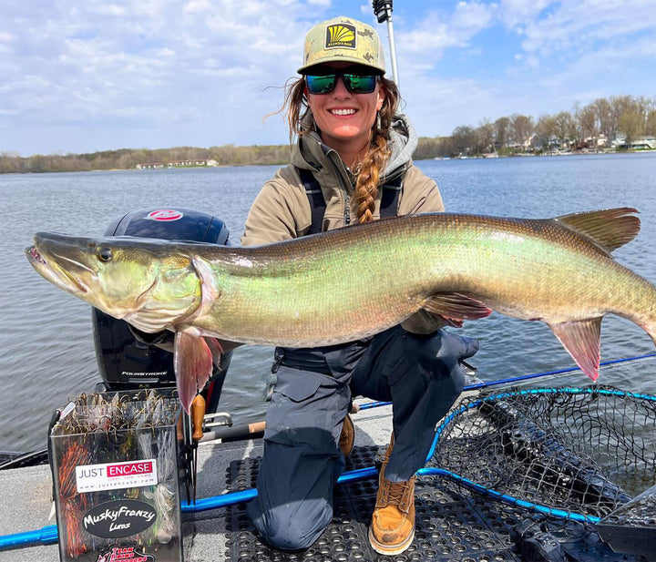 Christina Truppe is on a boat and smiling at the camera, while holding a large musky. There is a fly fishing net behind her lying on the boat. 