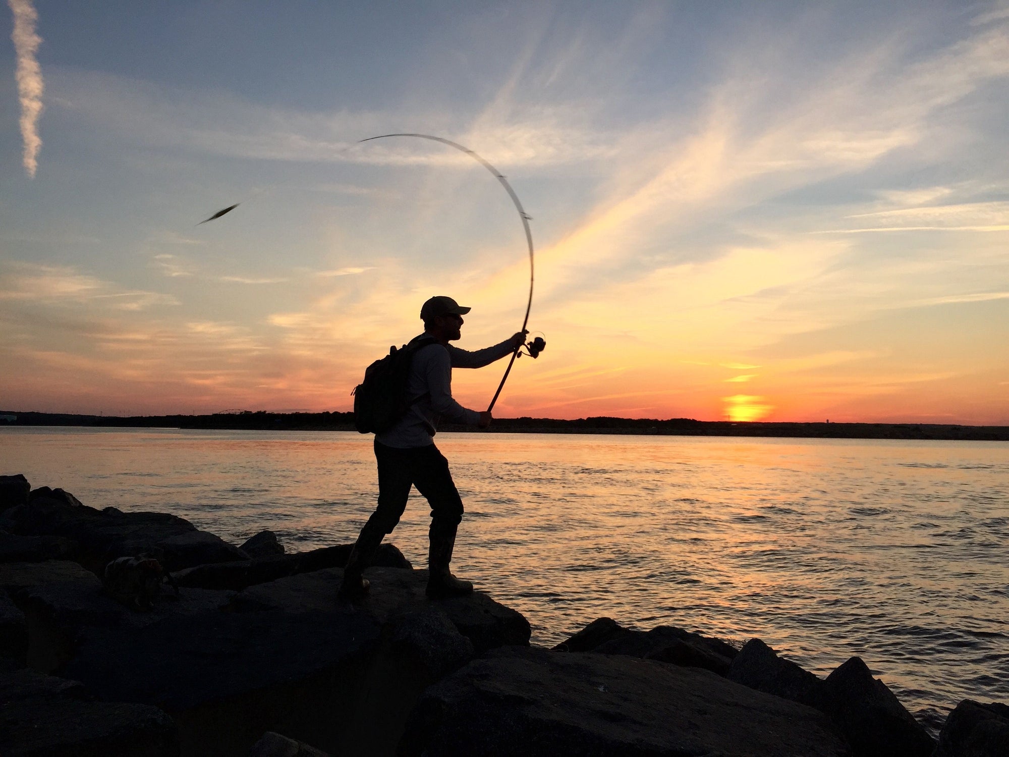 An individual is casting their fishing line with a sunset in the background. 