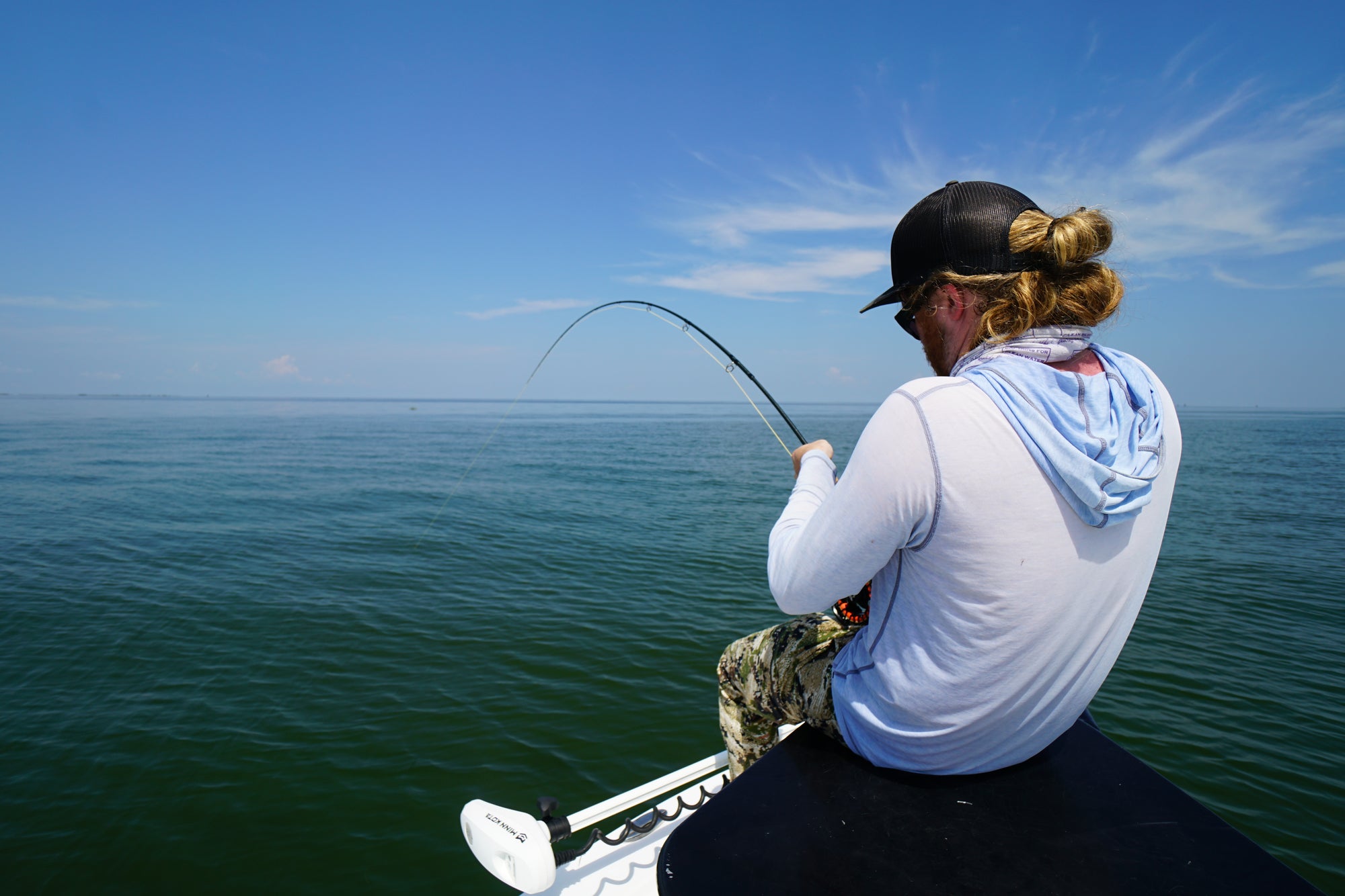 An angler is seated on a boat with his back towards the camera. He is reeling in his line. 